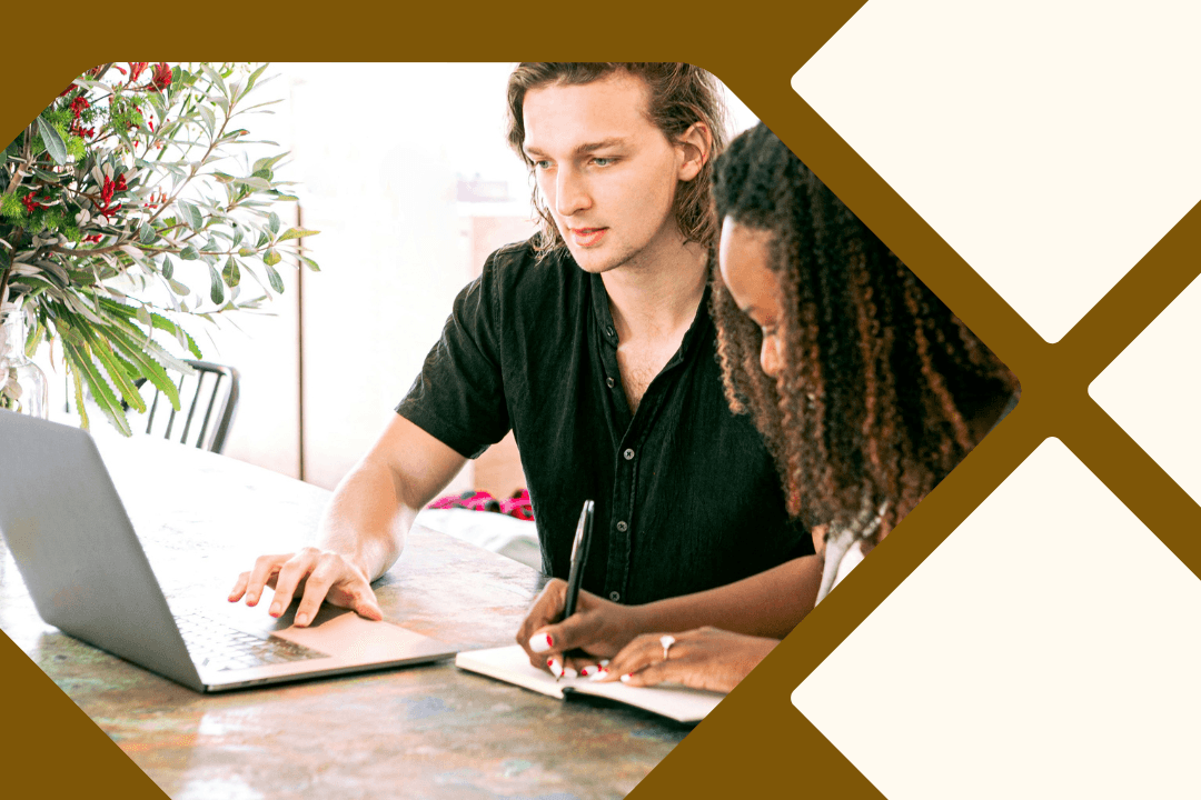 A man and a woman are sitting at a desk working on a laptop. The woman is also taking notes in a notebook on the desk.